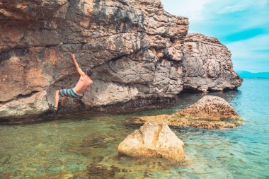Güçlü bir adam sabah vakti sahildeki kayalara tırmanıyor ve doğanın tadını çıkarıyor. Denize tırmanan. Yaz maceraları ve tatil konsepti. Adrenalin sporları.