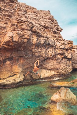 Güçlü bir adam sabah vakti sahildeki kayalara tırmanıyor ve doğanın tadını çıkarıyor. Denize tırmanan. Yaz maceraları ve tatil konsepti. Adrenalin sporları.