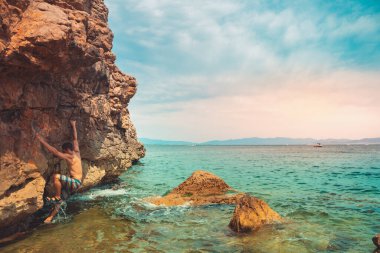 Güçlü bir adam sabah vakti sahildeki kayalara tırmanıyor ve doğanın tadını çıkarıyor. Denize tırmanan. Yaz maceraları ve tatil konsepti. Adrenalin sporları.