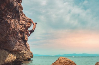 Güçlü bir adam sabah vakti sahildeki kayalara tırmanıyor ve doğanın tadını çıkarıyor. Denize tırmanan. Yaz maceraları ve tatil konsepti. Adrenalin sporları.