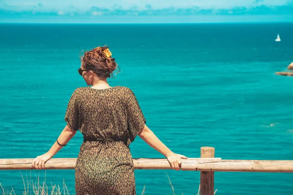 Beautiful woman standing and enjoy watching the sea view at balcony. Summer vibes. Adventure and travel female with leopard dress and sunglasses. Freedom, vacation and relax concept.