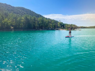 Yemek masasında güzel ve güçlü bir kadın gölde şeffaf suların tadını çıkarıyor. Tropik seyahat, seyahat tutkusu ve su aktivitesi konsepti. Paddle Surf su sporları. Yaz tatilleri.