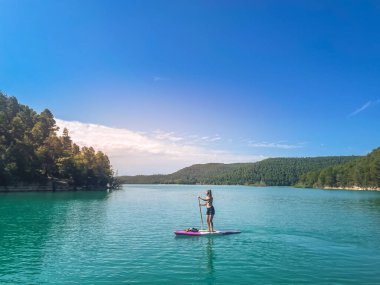 Yemek masasında güzel ve güçlü bir kadın gölde şeffaf suların tadını çıkarıyor. Tropik seyahat, seyahat tutkusu ve su aktivitesi konsepti. Paddle Surf su sporları. Yaz tatilleri.