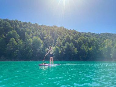 Yemek masasında güzel ve güçlü bir kadın gölde şeffaf suların tadını çıkarıyor. Tropik seyahat, seyahat tutkusu ve su aktivitesi konsepti. Paddle Surf su sporları. Yaz tatilleri.