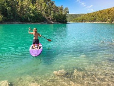 Yemek masasında güzel ve güçlü bir kadın gölde şeffaf suların tadını çıkarıyor. Tropik seyahat, seyahat tutkusu ve su aktivitesi konsepti. Paddle Surf su sporları. Yaz tatilleri.