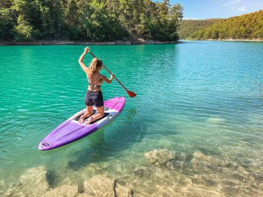 Yemek masasında güzel ve güçlü bir kadın gölde şeffaf suların tadını çıkarıyor. Tropik seyahat, seyahat tutkusu ve su aktivitesi konsepti. Paddle Surf su sporları. Yaz tatilleri.