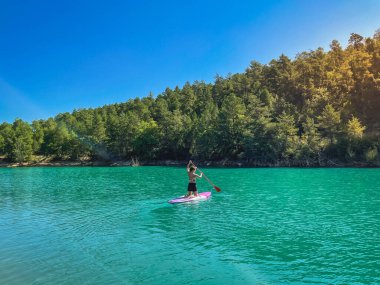 Yemek masasında güzel ve güçlü bir kadın gölde şeffaf suların tadını çıkarıyor. Tropik seyahat, seyahat tutkusu ve su aktivitesi konsepti. Paddle Surf su sporları. Yaz tatilleri.