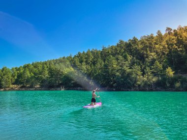 Yemek masasında güzel ve güçlü bir kadın gölde şeffaf suların tadını çıkarıyor. Tropik seyahat, seyahat tutkusu ve su aktivitesi konsepti. Paddle Surf su sporları. Yaz tatilleri.