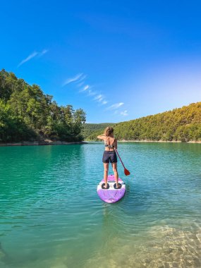 Yemek masasında güzel ve güçlü bir kadın gölde şeffaf suların tadını çıkarıyor. Tropik seyahat, seyahat tutkusu ve su aktivitesi konsepti. Paddle Surf su sporları. Yaz tatilleri.