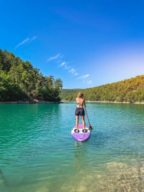 Yemek masasında güzel ve güçlü bir kadın gölde şeffaf suların tadını çıkarıyor. Tropik seyahat, seyahat tutkusu ve su aktivitesi konsepti. Paddle Surf su sporları. Yaz tatilleri.