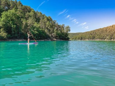 Yemek masasında güzel ve güçlü bir kadın gölde şeffaf suların tadını çıkarıyor. Tropik seyahat, seyahat tutkusu ve su aktivitesi konsepti. Paddle Surf su sporları. Yaz tatilleri.