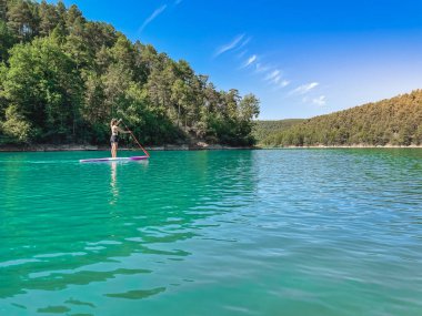 Yemek masasında güzel ve güçlü bir kadın gölde şeffaf suların tadını çıkarıyor. Tropik seyahat, seyahat tutkusu ve su aktivitesi konsepti. Paddle Surf su sporları. Yaz tatilleri.