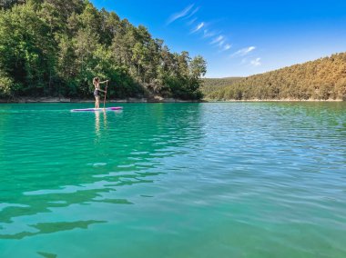 Yemek masasında güzel ve güçlü bir kadın gölde şeffaf suların tadını çıkarıyor. Tropik seyahat, seyahat tutkusu ve su aktivitesi konsepti. Paddle Surf su sporları. Yaz tatilleri.