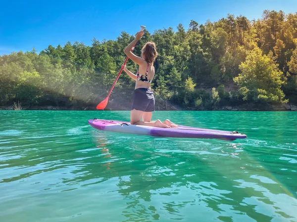 Yemek masasında güzel ve güçlü bir kadın gölde şeffaf suların tadını çıkarıyor. Tropik seyahat, seyahat tutkusu ve su aktivitesi konsepti. Paddle Surf su sporları. Yaz tatilleri.