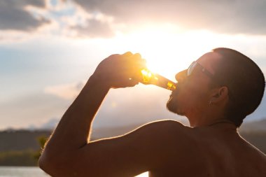 Genç adam gün batımında gölde eğleniyor. Kamp sandalyesinde oturup şişeden soğuk bira içmek. Tatiller, tatiller ve rahatlama konsepti.