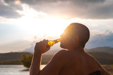 Genç adam gün batımında gölde eğleniyor. Kamp sandalyesinde oturup şişeden soğuk bira içmek. Tatiller, tatiller ve rahatlama konsepti.