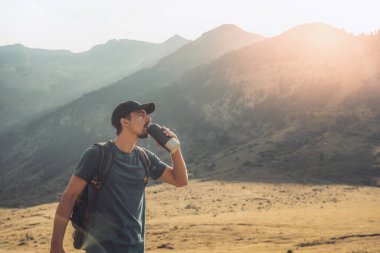Genç adam gün batımında muhteşem manzara manzaralı yeşil bir dağın tepesinde su içiyor. Cennet çim dağı. Spor ve sağlıklı aktiviteler. Su iç konsepti.