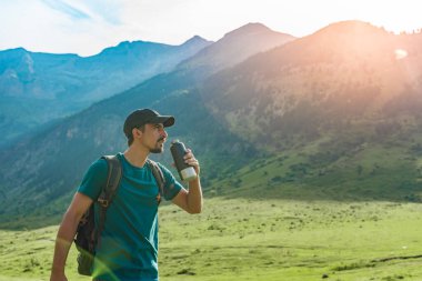 Genç adam gün batımında muhteşem manzara manzaralı yeşil bir dağın tepesinde su içiyor. Cennet çim dağı. Spor ve sağlıklı aktiviteler. Su iç konsepti.