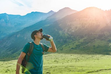 Genç adam gün batımında muhteşem manzara manzaralı yeşil bir dağın tepesinde su içiyor. Cennet çim dağı. Spor ve sağlıklı aktiviteler. Su iç konsepti.