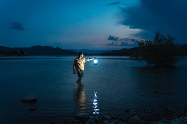 Gece vakti elinde fenerle göldeki suyun içindeki genç adam. Güneş ışığı, çocuğun güzel manzaraya bakışını gösterir. Karanlıkta tek başına. Antika bir fener gece yolu aydınlatıyor. Umut kavramı.