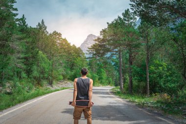 Genç hippi elinde havalı bir uzun tahta tutuyor. Güzel bir seyahat konsepti. Milenyum macera yolculuğu. Kaykay kaykay gezisi. Vahşi doğada spor.