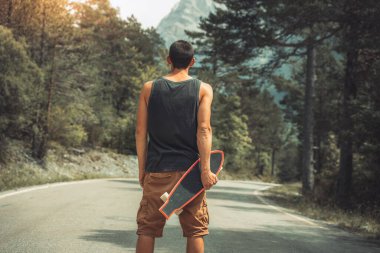 Genç hippi elinde havalı bir uzun tahta tutuyor. Güzel bir seyahat konsepti. Milenyum macera yolculuğu. Kaykay kaykay gezisi. Vahşi doğada spor.