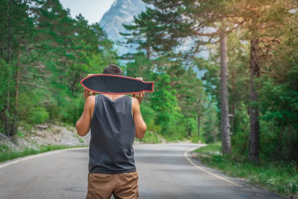Genç hippi elinde havalı bir uzun tahta tutuyor. Güzel bir seyahat konsepti. Milenyum macera yolculuğu. Kaykay kaykay gezisi. Vahşi doğada spor.