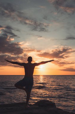 Formda bir genç adam gün batımında plajda yoga pozu veriyor. Meditasyon ve konsantrasyon duruşu, dinlenme gününün deniz tarafında, şafakta doğa tarafından çevrelenmiş..
