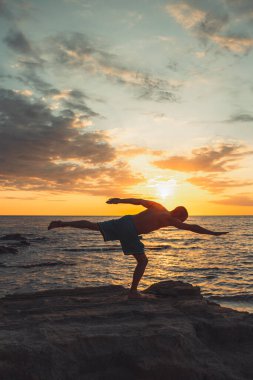 Formda bir genç adam gün batımında plajda yoga pozu veriyor. Meditasyon ve konsantrasyon duruşu, dinlenme gününün deniz tarafında, şafakta doğa tarafından çevrelenmiş..