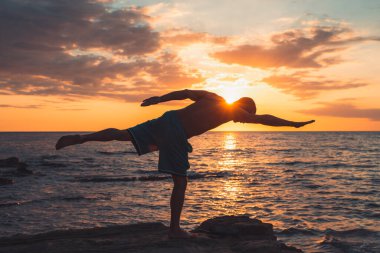 Formda bir genç adam gün batımında plajda yoga pozu veriyor. Meditasyon ve konsantrasyon duruşu, dinlenme gününün deniz tarafında, şafakta doğa tarafından çevrelenmiş..
