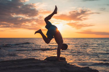 Formda bir genç adam gün batımında plajda yoga pozu veriyor. Meditasyon ve konsantrasyon duruşu, dinlenme gününün deniz tarafında, şafakta doğa tarafından çevrelenmiş..