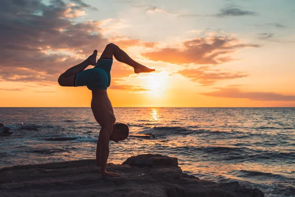 Formda bir genç adam gün batımında plajda yoga pozu veriyor. Meditasyon ve konsantrasyon duruşu, dinlenme gününün deniz tarafında, şafakta doğa tarafından çevrelenmiş..