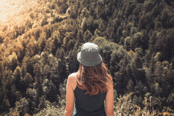 Young woman standing alone outdoors with wild forest mountains in background. Traveling lifestyle and survival concept rear view. Trekking with a green hat.