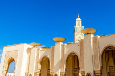 Kazablanka, Fas 'taki Hasan II Camii' nin güneşli dış galerisinde oyulmuş kemerler, çatı dekoratif kaseler ve minarenin uzak bir manzarası yer alıyor. Parlak mavi gökyüzü ışığı vurgular