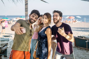 Gençler bir araya gelip sahilde selfie çekiyorlar. Akıllı telefon kullanan ve arka planda deniz varken eğlenen arkadaşlar.