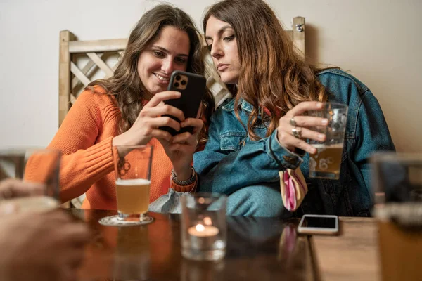 Two young women friends sharing smartphone screen drinking beers sitting at pubs table. Youth using alcohol and technology concept.