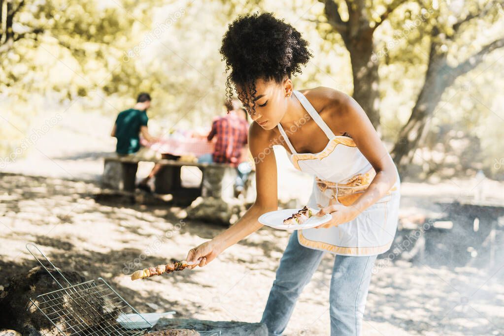 A young African-American girl picks up skewers from the barbecue while her friends, out of focus in the background, eat together on a Sunday outing.