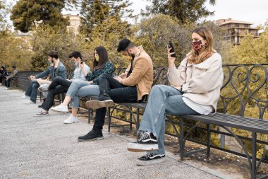 Koronavirüs kısıtlamaları nedeniyle parktaki akıllı telefonu kullanan gençlerin teknoloji kavramı covid-19 karantinası sırasında sosyal mesafeye saygılıdır..