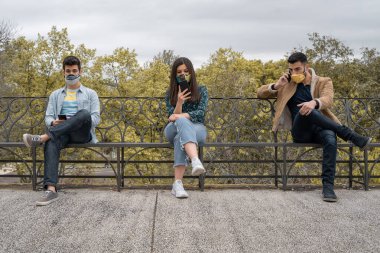 Milenyum kuşağı, akıllı telefon kullanan yüz maskesi takan gençler bir bankta oturmuş birbirlerine mesafeli oturuyorlar. Sosyal medya ve sosyal mesafe kavramı.