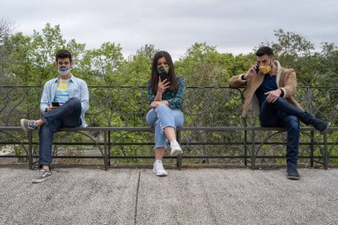 Koronavirüs kısıtlamaları nedeniyle parktaki akıllı telefonu kullanan gençlerin teknoloji kavramı covid-19 karantinası sırasında sosyal mesafeye saygılıdır..