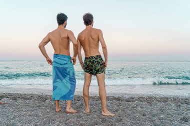 Arkadan sarılan iki genç adam sahilde ufku seyrediyor.