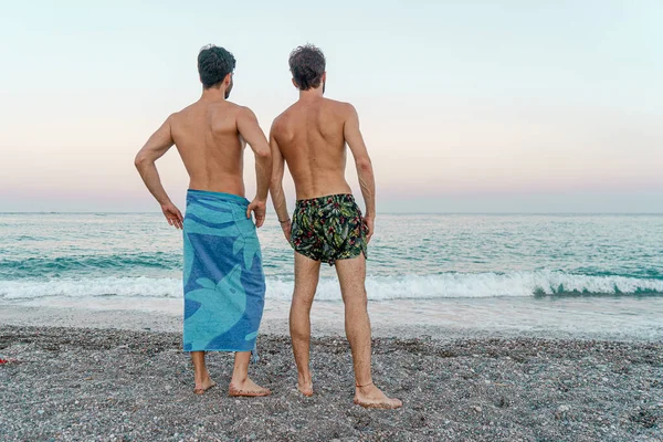 Arkadan sarılan iki genç adam sahilde ufku seyrediyor.