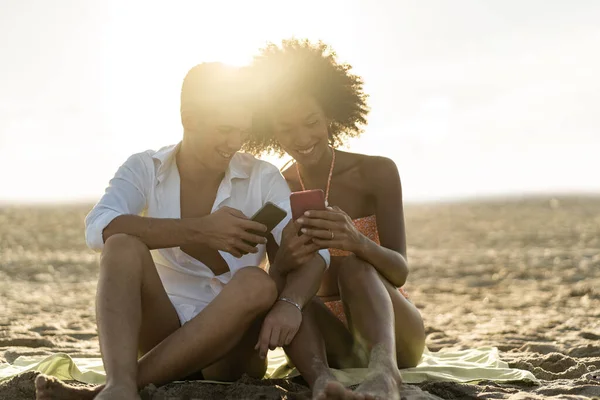 Interracial couple having fun watching social media content on smartphone screen sitting on the sand at the beach at sunset