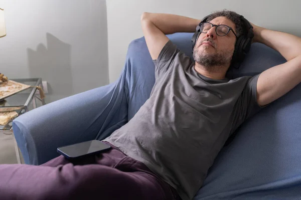 Orta yaşlı bir adam kanepede dinleniyor ve akıllı telefondan kablosuz kulaklıkla müzik dinliyor.