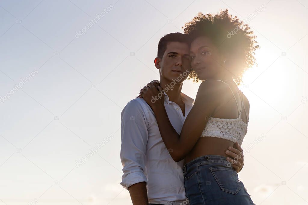 Portrait of mixed race couple of young lovers watching the camera with sun backlight against a blue sky.