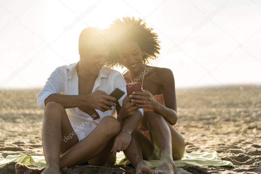 Interracial couple having fun watching social media content on smartphone screen sitting on the sand at the beach at sunset