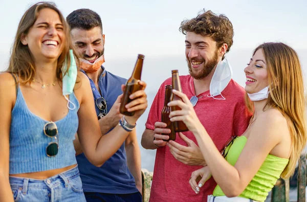 New normal lifestyle concept of young people hangout toasting with beer bottles outdoors during coronavirus breakdown wearing lowered face masks