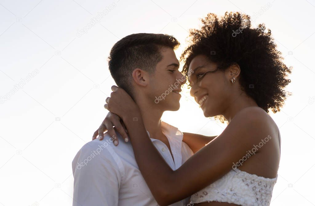 Multiracial couple with african american young woman and caucasian man hugging outdoor in a warm summer sunset. Tender romance and relationship interracial concept.