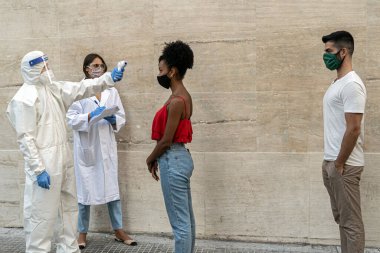 Coronavirus sıcaklık kontrol noktası. Bir görevli, genç bir Afro-Amerikalı kadının vücut ısısını ölçüyor. Diğer gençlerle aynı hizada, tabanca termometresiyle..