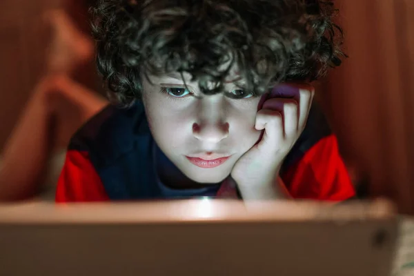 ビデオコンテンツを見たり タブレットコンピュータを持っているビデオゲームを夜の間に足で遊んでいる小さな男の子は 彼の顔は色のついたスクリーンライトで照らされています インターネット技術に依存している子供は — ストック写真
